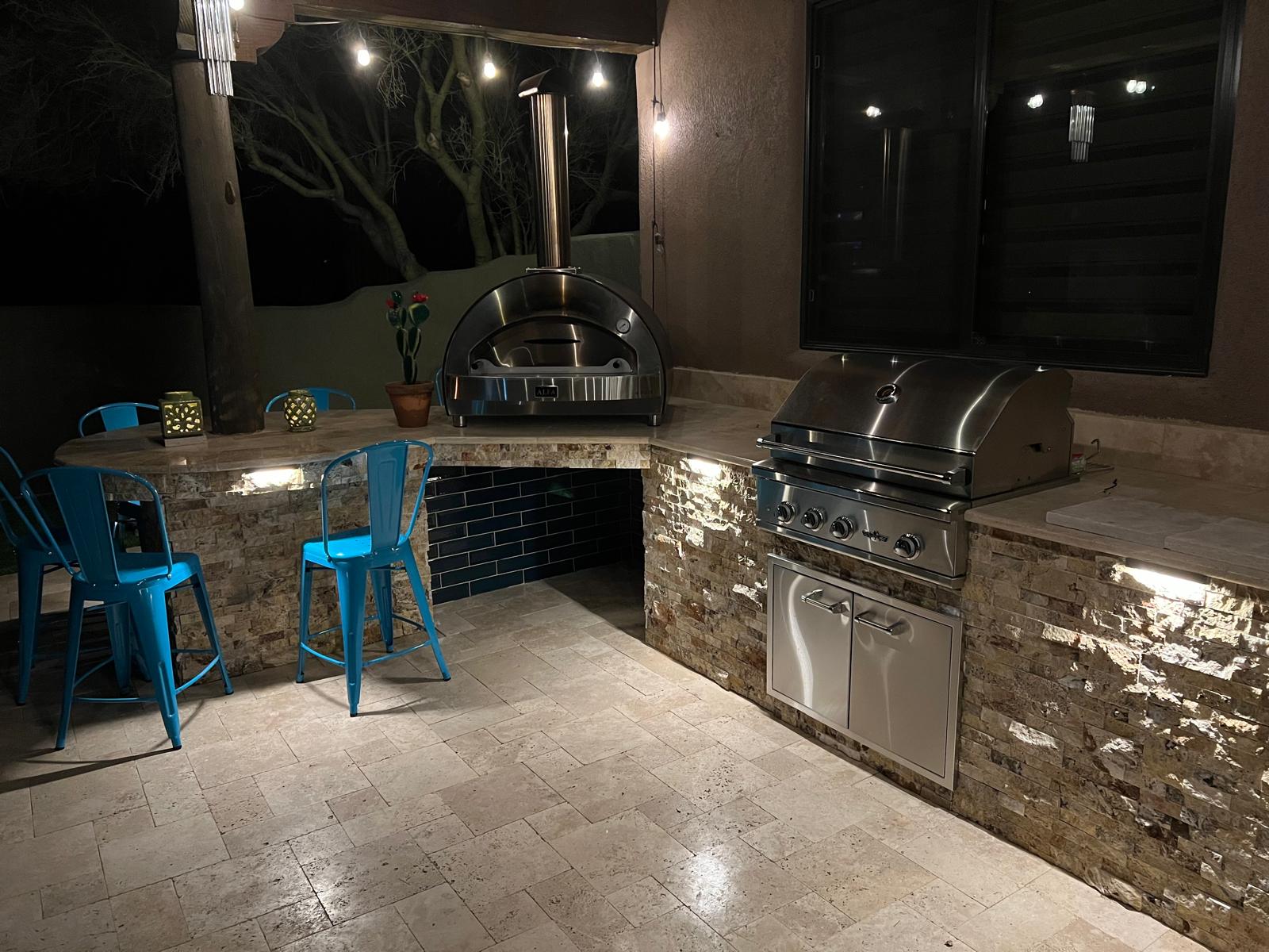 Illuminated outdoor kitchen with built-in grill, pizza oven and bar seating at night in Arizona