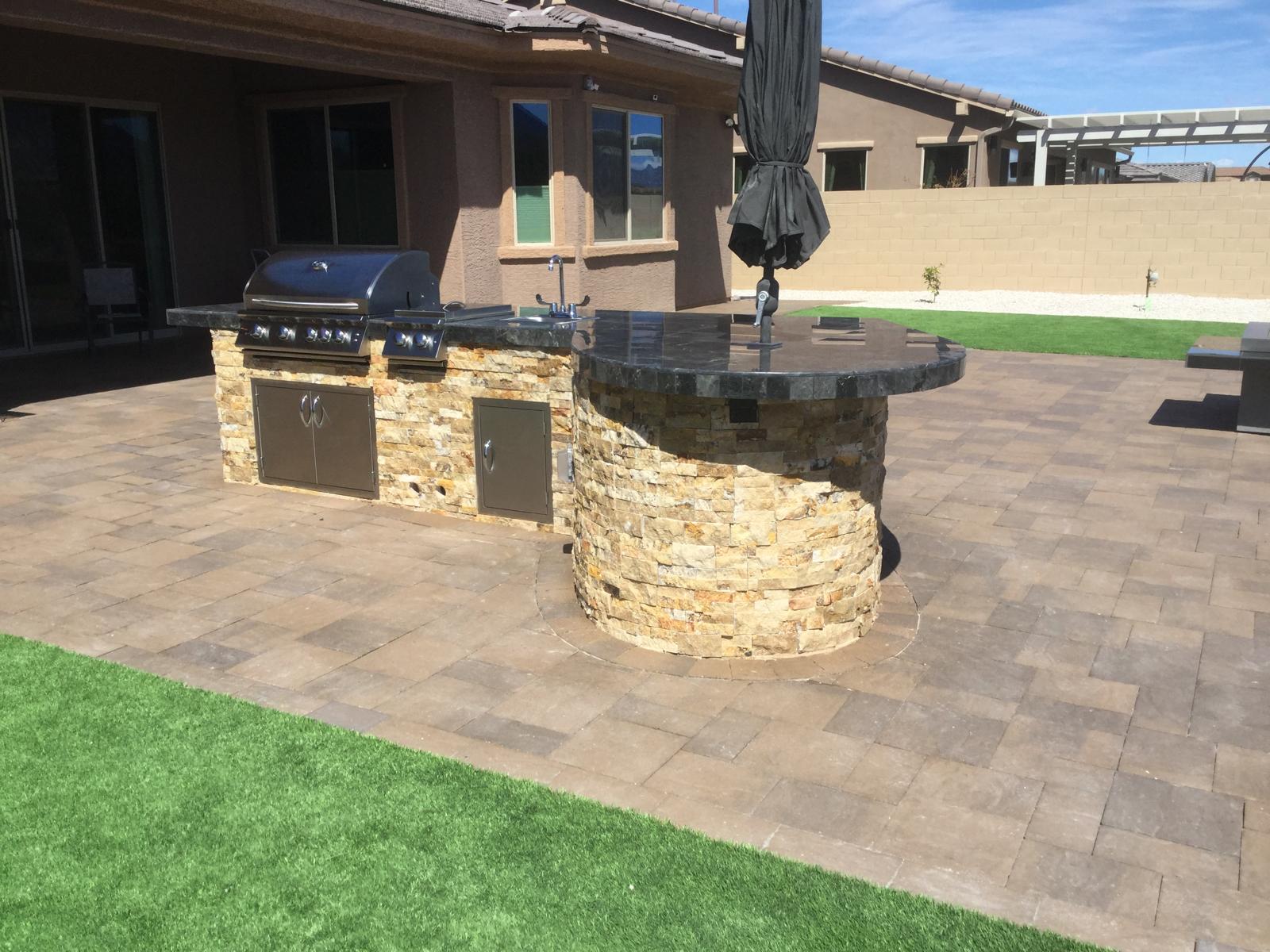 Curved stone outdoor kitchen and bar built on a paver patio with artificial grass in Arizona